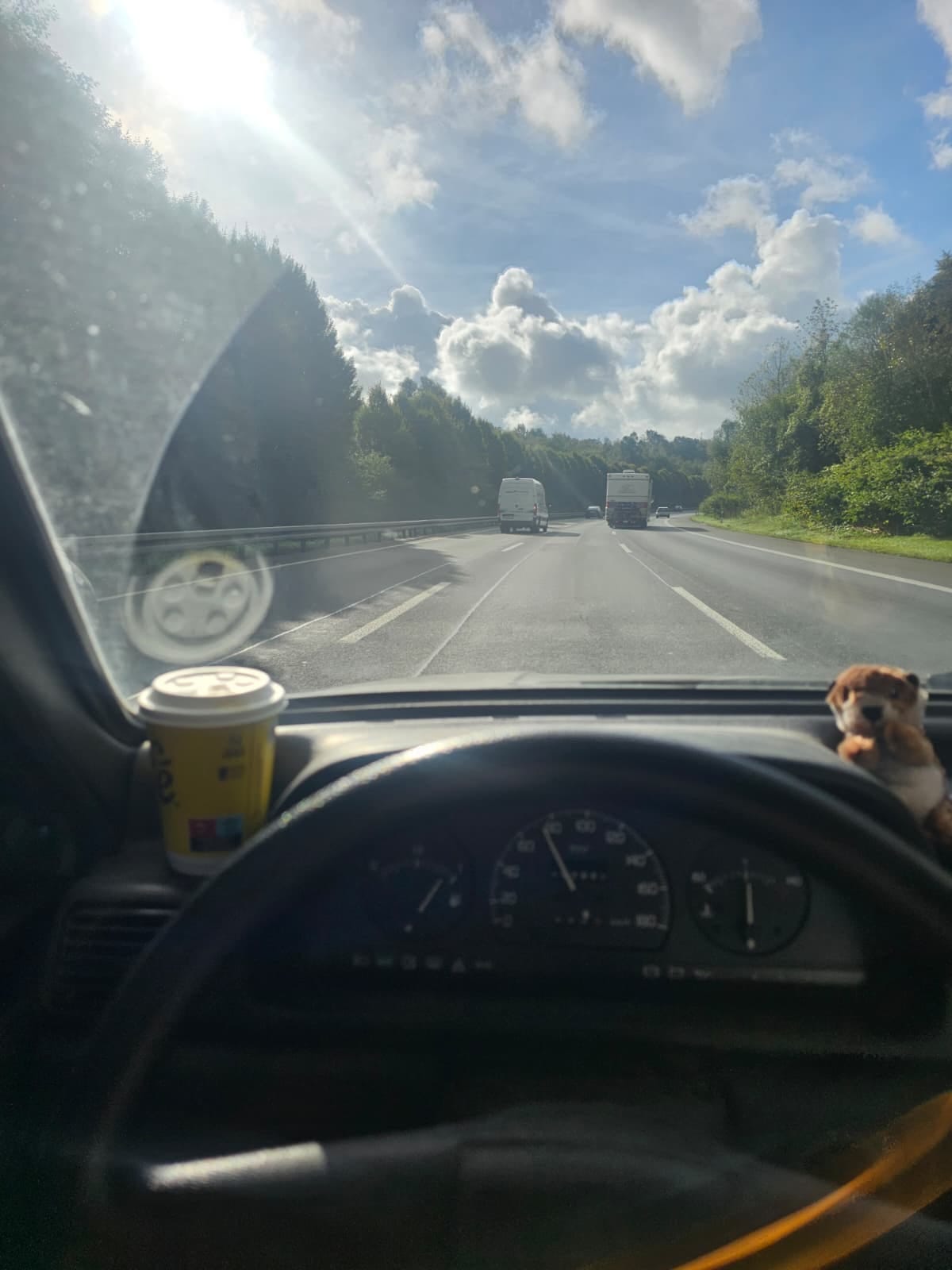 Blick übers Lenkrad auf die sonnige Autobahn, Kaffeebecher und Kuscheltier auf dem Armaturenbrett