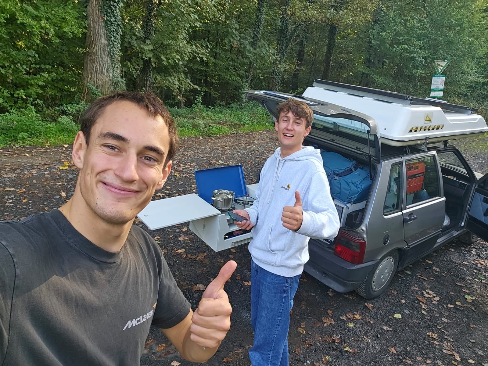Selfie im Wald: Daumen hoch vor dem Uno, Campingkocher in der Heckküche, Kofferraum voller Rucksäcke