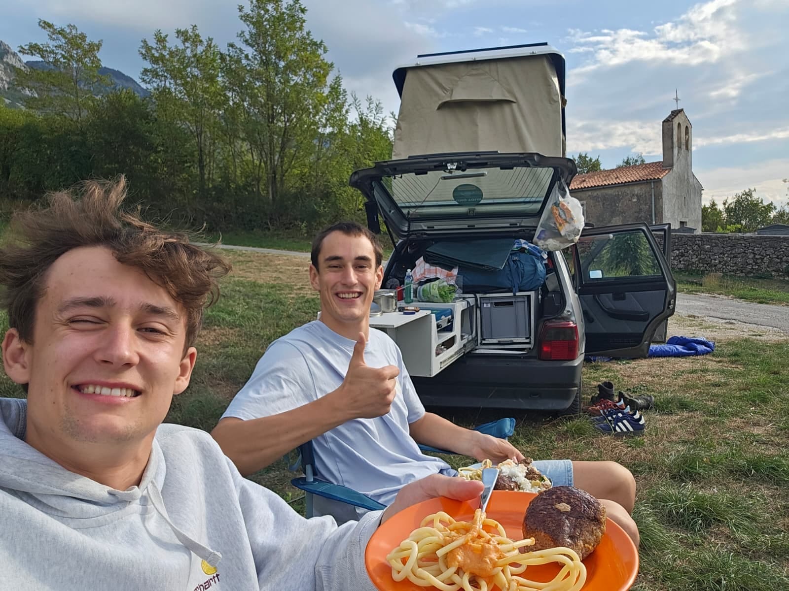 Mittagessen im Campingstuhl vor dem Uno mit offener Heckklappe und Dachzelt, im Hintergrund eine kleine Steinkapelle