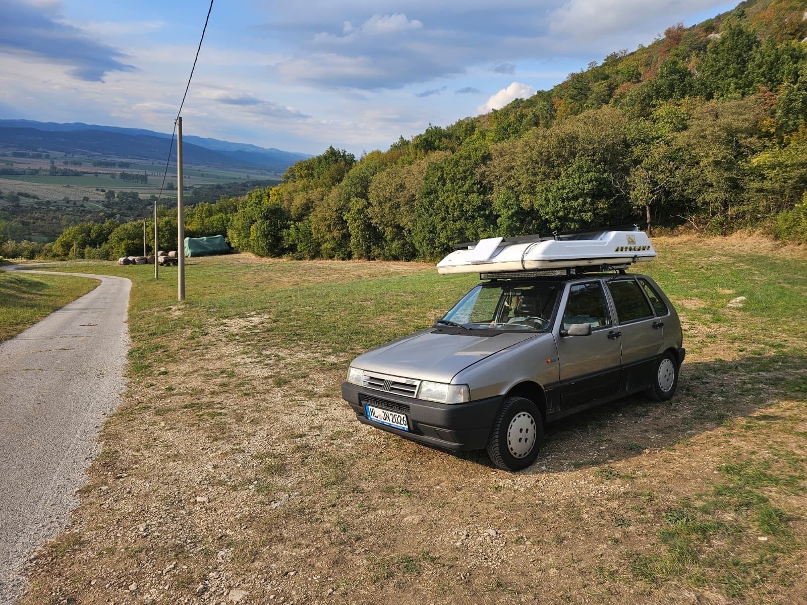 Unser silberner Fiat Uno mit weißem Dachzelt auf einer Wiese am Hang, dahinter Wald und ein weites Tal